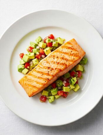 Grilled salmon fillet topped with fresh avocado salsa, garnished with cilantro on white plate